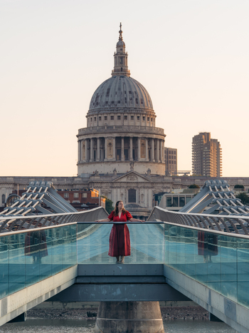 View of a woman in a vibrant red dress poised on the sleek Millennium Bridge with the majestic St. Paul s Cathedral gleaming in the golden light London England United Kingdom. View of a woman in a vibrant red dress poised on the sleek Millennium Bridge with the majestic St. Paul s Cathedral gleaming in the golden light London England United Kingdom.