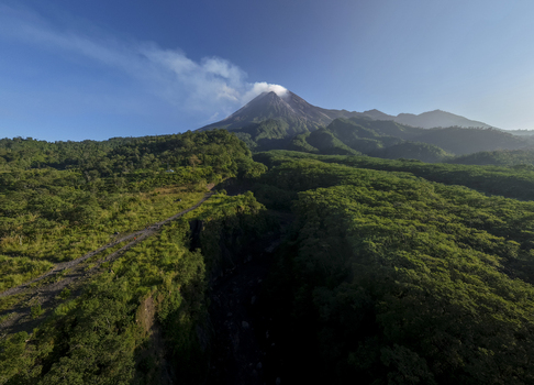 Aerial view of Bunker Kaliadem Merapi and the smoking peak of Mount Merapi volcano surrounded by lush green forests and rolling hills Kecamatan Pakem Daerah Istimewa Yogya Indonesia. Aerial view of Bunker Kaliadem Merapi and the smoking peak of Mount Merapi volcano surrounded by lush green forests and rolling hills Kecamatan Pakem Daerah Istimewa Yogya Indonesia.
