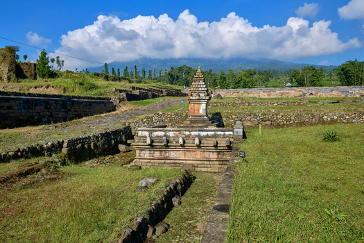 View of the ancient Situs Liyangan temple ruins with terraced stone walls and Mount Sindoro under a blue sky in Temanggung Central Java Indonesia.