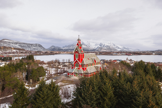 Aerial view of the vibrant red Buksnes Church stands proudly amidst evergreen trees against the backdrop of snow capped mountains Buksnes Kirke Nordland Norway. Aerial view of the vibrant red Buksnes Church stands proudly amidst evergreen trees against the backdrop of snow capped mountains Buksnes Kirke Nordland Norway.