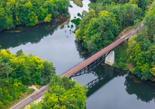 Aerial view of the Historic Railway Truss Bridge in Rutki spanning a calm river surrounded by dense green forest foliage in Rutki Pomeranian Voivodeship Poland. Aerial view of the Historic Railway Truss Bridge in Rutki spanning a calm river surrounded by dense green forest foliage in Rutki Pomeranian Voivodeship Poland.