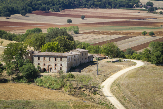 View of a winding road leading to an aged stone building amidst a rural landscape of varied textures and colors in Monteriggioni Tuscany Italy. View of a winding road leading to an aged stone building amidst a rural landscape of varied textures and colors in Monteriggioni Tuscany Italy.