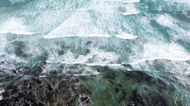 Aerial view of turquoise ocean waves with white foam crashing against dark rocky cliffs at Constantine Bay in Padstow Cornwall United Kingdom. Aerial view of turquoise ocean waves with white foam crashing against dark rocky cliffs at Constantine Bay in Padstow Cornwall United Kingdom.