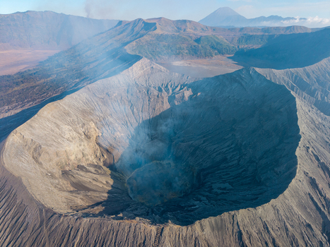 Aerial view of Mount Bromo crater with volcanic smoke rugged ash covered slopes and surrounding mountain range under clear sky in Tosari East Java Indonesia.