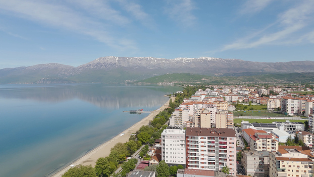 Aerial view of Pogradec city along the shore of Lake Ohrid featuring residential buildings a sandy beach and snow capped mountains under a clear blue sky. Pogradec Korce County Albania. Aerial view of Pogradec city along the shore of Lake Ohrid featuring residential buildings a sandy beach and snow capped mountains under a clear blue sky. Pogradec Korce County Albania.