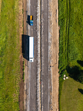 Aerial view of E67 Via Baltica road construction with trucks driving on the unfinished surface surrounded by green fields in Lielvarzi Kekava Municipality Latvia. Aerial view of E67 Via Baltica road construction with trucks driving on the unfinished surface surrounded by green fields in Lielvarzi Kekava Municipality Latvia.