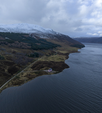 Aerial view of The Pink House on the shore of Loch Glass surrounded by rugged hills and snow capped mountains in Dingwall Scotland United Kingdom. Aerial view of The Pink House on the shore of Loch Glass surrounded by rugged hills and snow capped mountains in Dingwall Scotland United Kingdom.