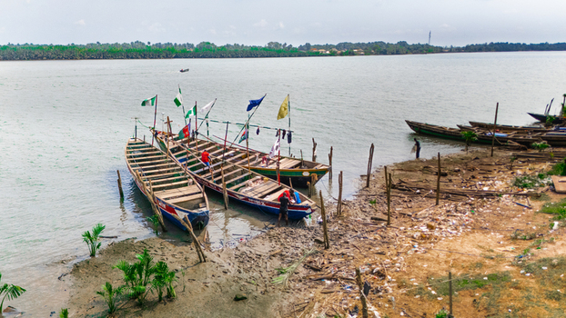 Aerial view of traditional wooden boats docked along the muddy riverbank with fishermen working nearby under a clear sky in Minima Rivers Nigeria.