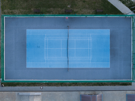 Aerial view of a blue tennis court with white lines and a net surrounded by a green fence in Poland.