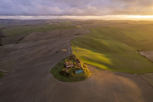 Aerial view of a villa with a swimming pool situated on a hill surrounded by the rolling landscape of the Crete Senesi at sunset in Asciano Tuscany Italy.