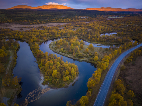 Aerial view of River Ljusnan winding through autumn forests with golden sunrise light on distant mountains and a road alongside the water in Bruksvallarna Jamtland County Sweden.