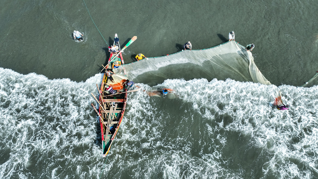 Aerial view of fishermen hauling in nets from a vibrant wooden boat amidst the churning foamy waves along the shore Cox s Bazar Chittagong Division Bangladesh. Aerial view of fishermen hauling in nets from a vibrant wooden boat amidst the churning foamy waves along the shore Cox s Bazar Chittagong Division Bangladesh.