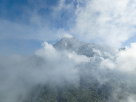 Aerial view of the majestic peak of Mount Merapi partially covered by thick white clouds under a clear blue sky in Kecamatan Cangkringan Daerah Istimewa Yogya Indonesia. Aerial view of the majestic peak of Mount Merapi partially covered by thick white clouds under a clear blue sky in Kecamatan Cangkringan Daerah Istimewa Yogya Indonesia.