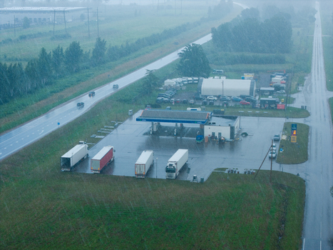 Cenu pagasts Latvia 04 July 2026 Aerial view of Virsi Brankstari gas station in heavy rain with parked semi trucks on a wet asphalt lot surrounded by green fields and a highway.