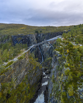 Aerial view of Voringfossen waterfall and the modern pedestrian bridge spanning a deep rocky canyon with lush vegetation Eidfjord Municipality Vestland Norway.