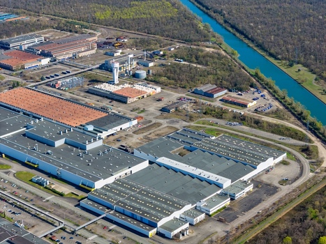 Aerial view of industrial buildings sprawling under a blue sky with a canal snaking nearby contrasting with the green trees Sausheim Grand Est France.