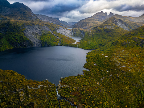 Aerial view of Stuvdalsvatnet lake surrounded by rugged mountains lush green vegetation and rocky terrain under a cloudy sky in Moskenes Nordland Norway. Aerial view of Stuvdalsvatnet lake surrounded by rugged mountains lush green vegetation and rocky terrain under a cloudy sky in Moskenes Nordland Norway.