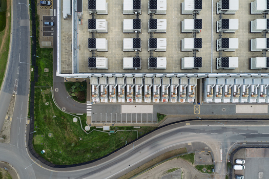 Aerial view of a large data center facility featuring numerous rooftop cooling units a paved parking area and surrounding access roads in the United Kingdom.