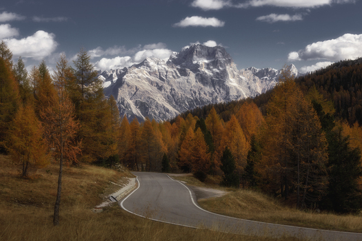 View of the Dolomites mountains towering over a winding road flanked by vibrant autumn larch trees in Rocca Pietore Veneto Italy. View of the Dolomites mountains towering over a winding road flanked by vibrant autumn larch trees in Rocca Pietore Veneto Italy.