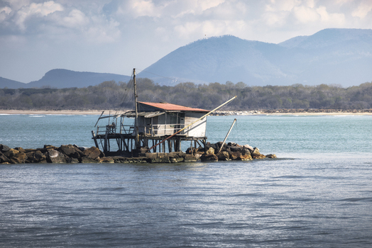 View of a traditional wooden fishing hut on stilts along a rocky breakwater with mountains in the background under a cloudy sky in Marina di Pisa Tuscany Italy.