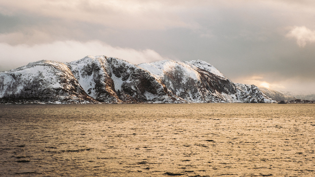 View of the sun reflecting off the water in front of snow capped mountains under a cloudy sky Brensholmen Troms Norway. View of the sun reflecting off the water in front of snow capped mountains under a cloudy sky Brensholmen Troms Norway.