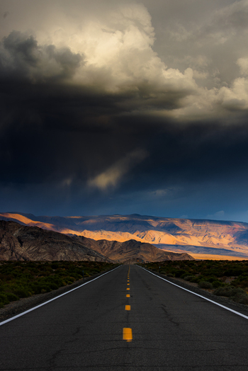 View of Death Valley National Park with a straight road leading towards mountains under dramatic dark storm clouds and golden light in the United States.