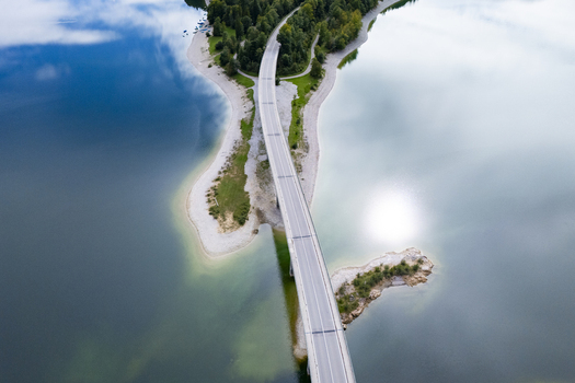 Aerial view of a bridge cutting through the shimmering Sylvensteinsee lake connecting forested peninsulas under a vast sky Sylvensteinspeicher Bayern Germany. Aerial view of a bridge cutting through the shimmering Sylvensteinsee lake connecting forested peninsulas under a vast sky Sylvensteinspeicher Bayern Germany.