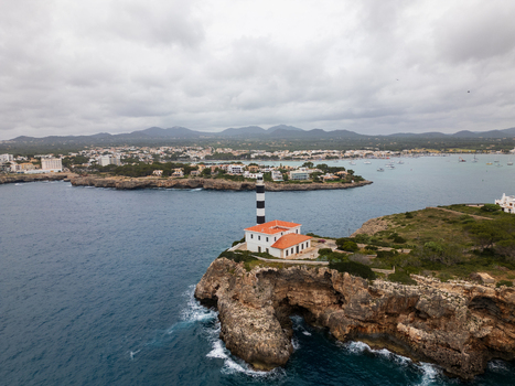 Aerial view of Far de Portocolom lighthouse on a rocky cliff overlooking the Mediterranean Sea with the town and mountains in the background Portocolom Balearic Islands Spain. Aerial view of Far de Portocolom lighthouse on a rocky cliff overlooking the Mediterranean Sea with the town and mountains in the background Portocolom Balearic Islands Spain.