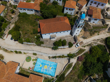 Aerial view of a traditional church with a blue dome and bell tower next to a bright blue basketball court and red roofed houses in Dhermi Vlore County Albania. Aerial view of a traditional church with a blue dome and bell tower next to a bright blue basketball court and red roofed houses in Dhermi Vlore County Albania.