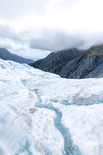 View of glacial ice carved with turquoise streams under a brooding sky meets rugged mountain peaks in Franz Josef Glacier West Coast Region New Zealand. View of glacial ice carved with turquoise streams under a brooding sky meets rugged mountain peaks in Franz Josef Glacier West Coast Region New Zealand.