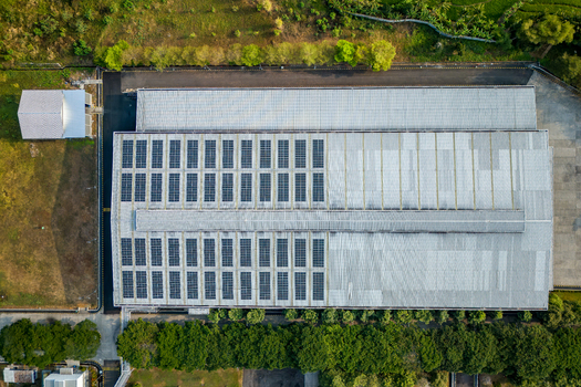 Aerial view of a large industrial warehouse roof featuring extensive solar panel arrays surrounded by green trees and paved areas in Semarang Regency Central Java Indonesia. Aerial view of a large industrial warehouse roof featuring extensive solar panel arrays surrounded by green trees and paved areas in Semarang Regency Central Java Indonesia.
