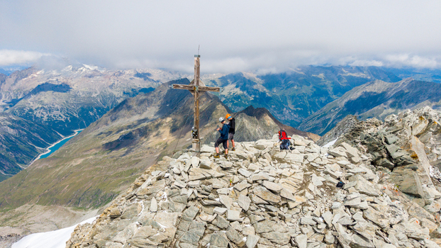 Aerial view of Grosser Moseler summit with a wooden cross two hikers on the rocky peak and a distant blue reservoir in the valley below Schwaz District Tyrol Austria. Aerial view of Grosser Moseler summit with a wooden cross two hikers on the rocky peak and a distant blue reservoir in the valley below Schwaz District Tyrol Austria.