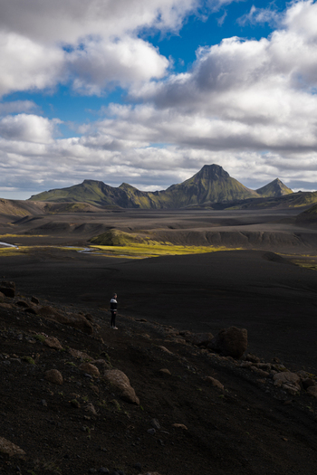 View of a lone figure stands amidst the stark black volcanic landscape under a sky dotted with clouds framed by distant mountains Skaftárhreppur Iceland. View of a lone figure stands amidst the stark black volcanic landscape under a sky dotted with clouds framed by distant mountains Skaftárhreppur Iceland.