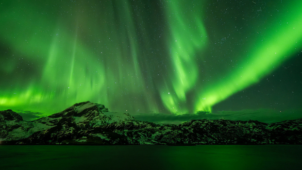View of the vibrant green aurora borealis illuminating the night sky above the snow capped mountains and dark waters in Leknes Lofoten Norway.