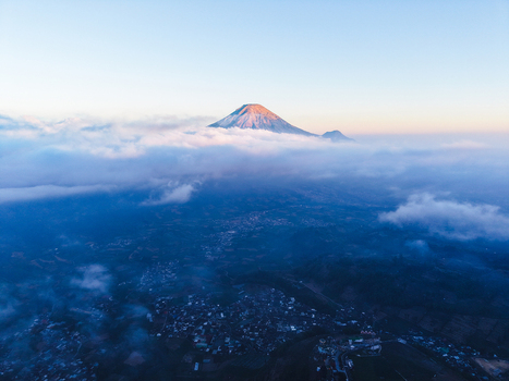 Aerial view of the Dieng Plateau mountain peak rising above a sea of clouds at sunrise with golden light on the summit and a village visible in the valley below in Indonesia.