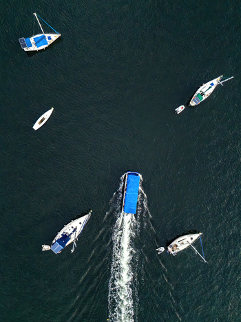 Aerial view of a blue motorboat cruising and leaving a long white wake while passing by several white sailboats anchored on the dark rippling surface of the water. Aerial view of a blue motorboat cruising and leaving a long white wake while passing by several white sailboats anchored on the dark rippling surface of the water.