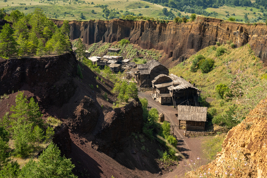 View of rustic wooden structures nestled within the steep dark red volcanic walls of the Racos scoria quarry surrounded by pine trees and grassy slopes in Brasov Brasov Romania.