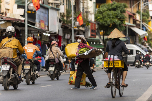 Hanoi Vietnam 11 November 2025 View of a street vendor wearing a conical hat crossing a busy street filled with motorbikes and bicycles in the historic Old Quarter.