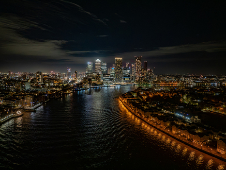 Aerial view of the illuminated Canary Wharf skyline at night with the River Thames reflecting city lights modern skyscrapers and urban architecture in London England United Kingdom. Aerial view of the illuminated Canary Wharf skyline at night with the River Thames reflecting city lights modern skyscrapers and urban architecture in London England United Kingdom.