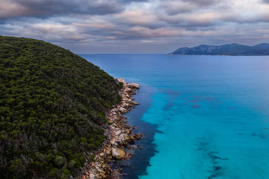 Aerial view of Bettys Beach featuring turquoise waters rocky coastline and dense green forest under a cloudy sky in Albany WA Australia. Aerial view of Bettys Beach featuring turquoise waters rocky coastline and dense green forest under a cloudy sky in Albany WA Australia.
