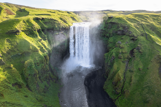 Aerial view of Skogafoss waterfall cascading down a steep cliff surrounded by lush green hills and a winding river in Rangarthing eystra Iceland.