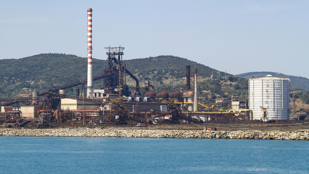 View of the industrial complex with a towering chimney juxtaposed against the serene sea and distant hills creating a striking contrast Piombino Tuscany Italy. View of the industrial complex with a towering chimney juxtaposed against the serene sea and distant hills creating a striking contrast Piombino Tuscany Italy.