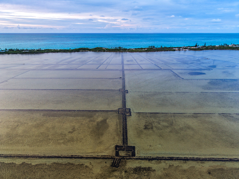 Aerial view of Salt Pond with geometric salt pans and a narrow causeway leading toward the turquoise ocean under a cloudy sky in Exuma Exuma The Bahamas.