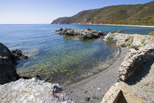 View of crystal clear turquoise water meeting a dark pebbled shore under a vast blue sky framed by rocky outcrops and distant green hills Lacona Elba Italy. View of crystal clear turquoise water meeting a dark pebbled shore under a vast blue sky framed by rocky outcrops and distant green hills Lacona Elba Italy.