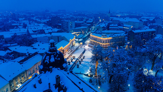 Aerial view of the snow covered town square and historic buildings illuminated by warm lights during the blue hour in Sremska Mitrovica Vojvodina Serbia.