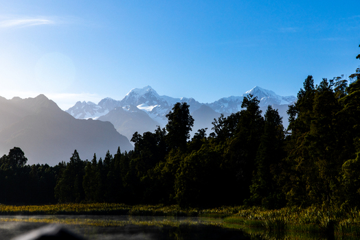 View of dark trees reflecting in the calm waters beneath snow capped mountains under a clear blue sky creating a tranquil scene Lake Matheson West Coast Region New Zealand. View of dark trees reflecting in the calm waters beneath snow capped mountains under a clear blue sky creating a tranquil scene Lake Matheson West Coast Region New Zealand.