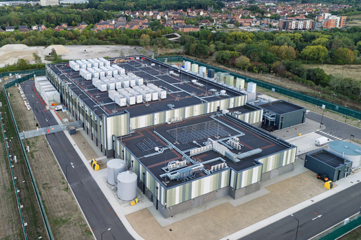 Aerial view of a large industrial data center with rooftop HVAC units and cooling infrastructure surrounded by a secure perimeter fence and green landscape in the United Kingdom. Aerial view of a large industrial data center with rooftop HVAC units and cooling infrastructure surrounded by a secure perimeter fence and green landscape in the United Kingdom.