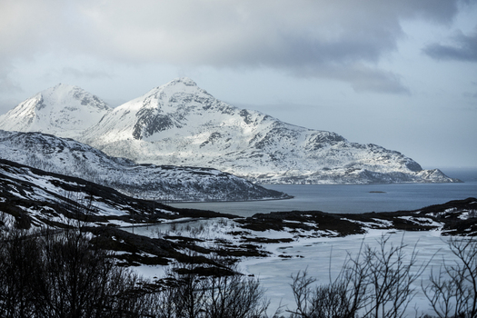 View of snow covered mountain peaks overlooking a calm fjord under a dramatic cloudy sky in Kvaloya Troms Norway. View of snow covered mountain peaks overlooking a calm fjord under a dramatic cloudy sky in Kvaloya Troms Norway.