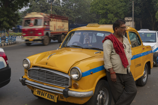 View of a man with a red cloth draped over his shoulder leans against a vintage yellow taxi a blur of a red truck speeding by in the background Kolkata West Bengal India. View of a man with a red cloth draped over his shoulder leans against a vintage yellow taxi a blur of a red truck speeding by in the background Kolkata West Bengal India.