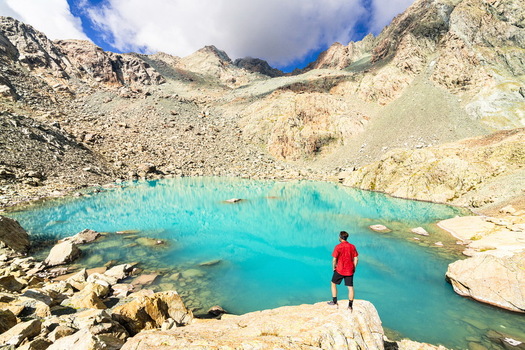 View of a man in a red shirt standing on a rock overlooking a turquoise alpine lake surrounded by rugged mountain peaks and rocky slopes in Chiesa in Valmalenco Lombardy Italy. View of a man in a red shirt standing on a rock overlooking a turquoise alpine lake surrounded by rugged mountain peaks and rocky slopes in Chiesa in Valmalenco Lombardy Italy.